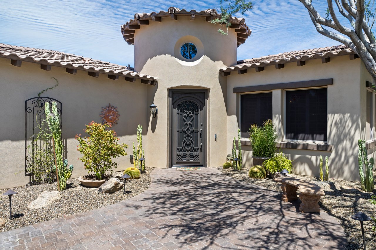 Front entrance of a stucco house with a tiled roof, arched tower detail, iron security door, desert landscaping, and brick paver walkway.