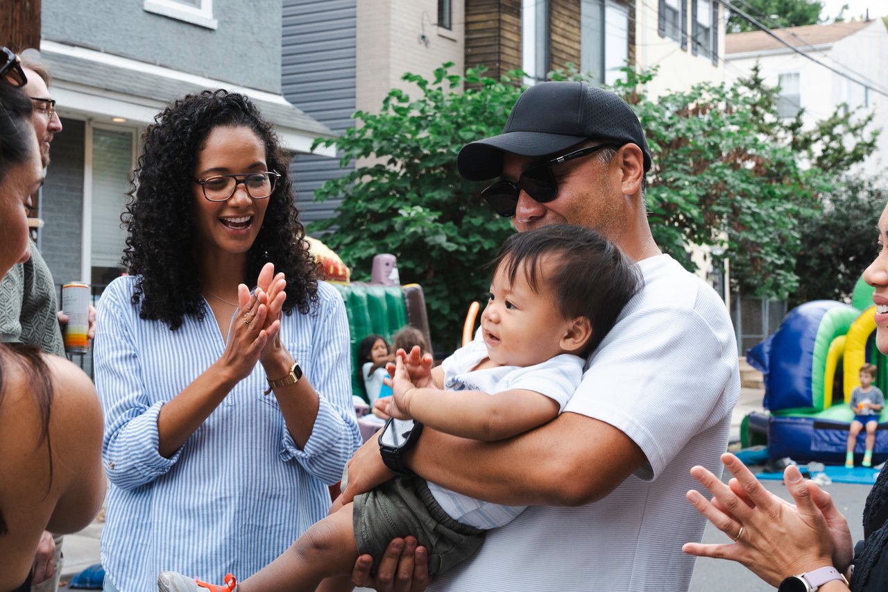 Neighbors celebrate at a block party as a woman claps and a man holds a baby.