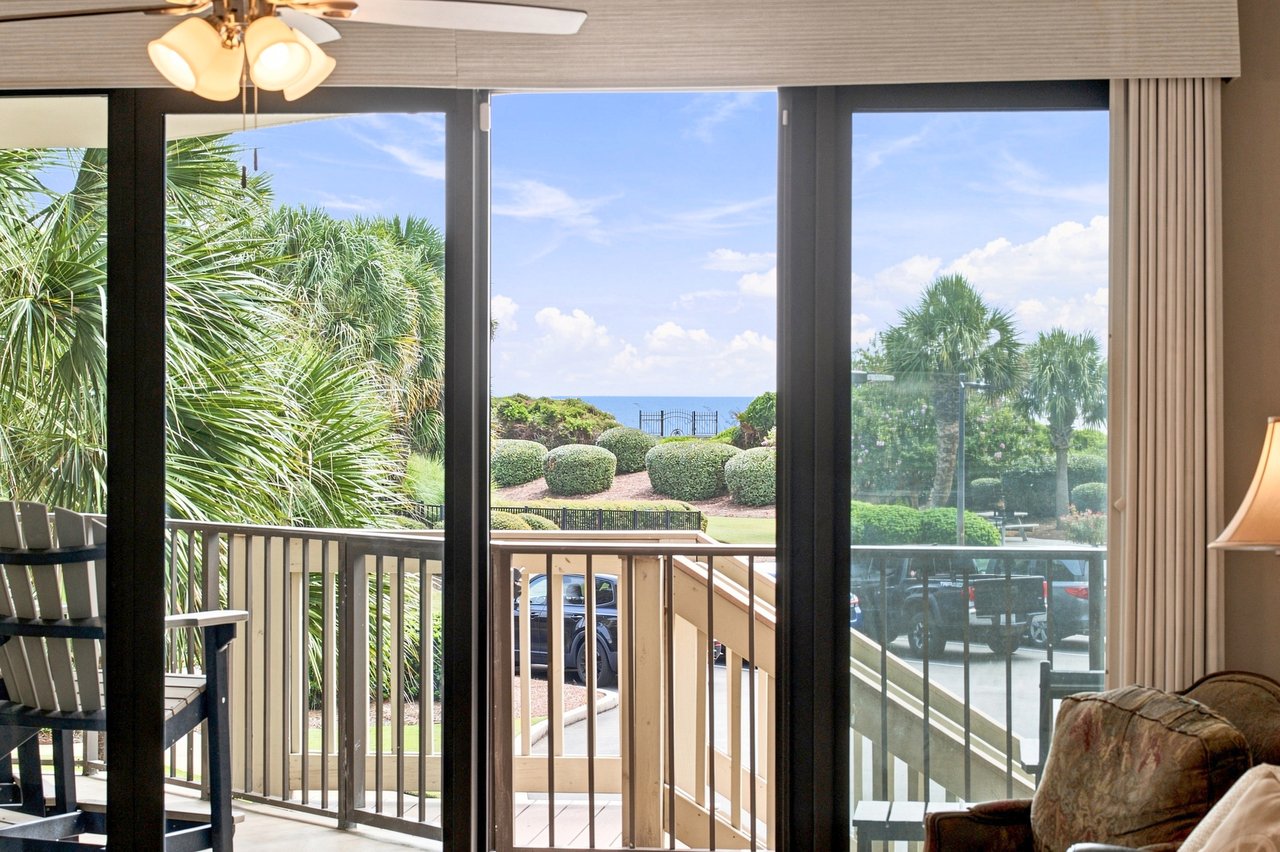 Living room opens to a balcony with palm trees, landscaped grounds, and a glimpse of the ocean.