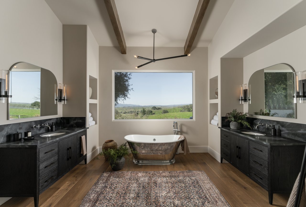 Bathroom with a freestanding tub, matching dark vanities, and countryside views through a large window.