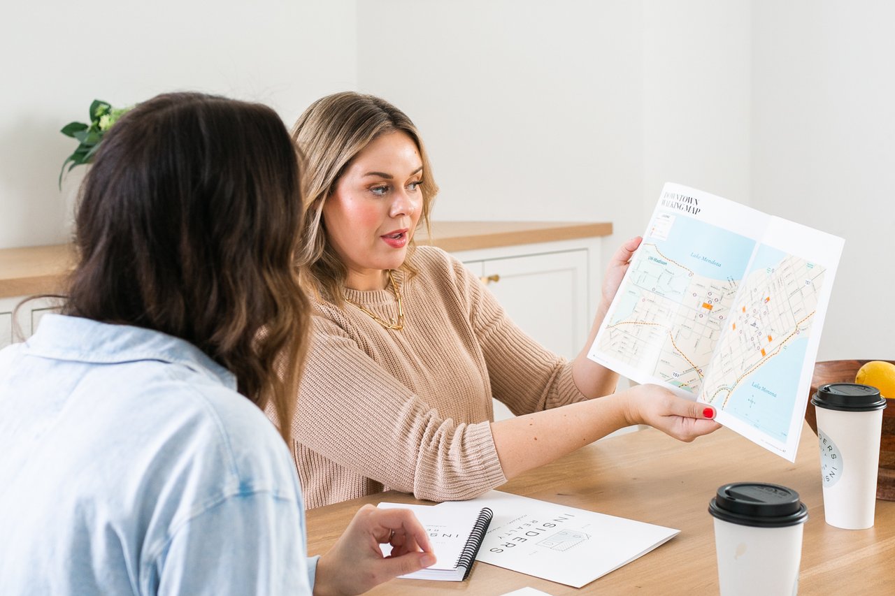 Cait Berry of Insiders Realty explaining a downtown Madison map to a buyer at a kitchen table, gesturing to the locations of Lake Mendota, Lake Monona, UW-Madison, and Highway 151, discussing how the geography connects to the Middleton commute and other Madison neighborhood options.
