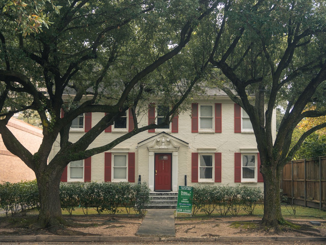 White brick house with red shutters and front door framed by large mature trees.