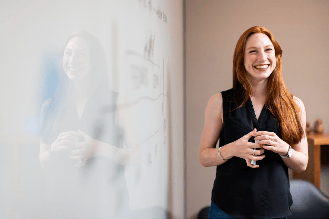 Smiling red-haired woman standing beside a whiteboard.