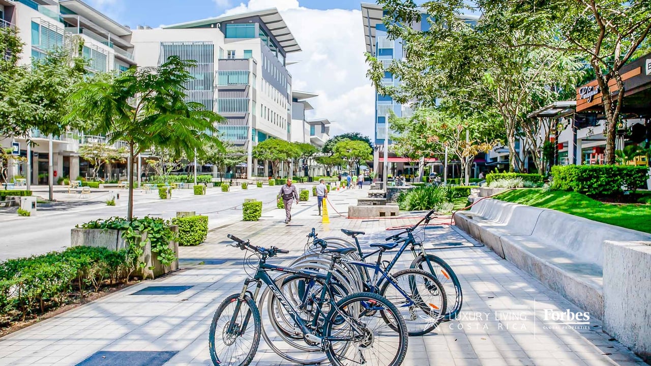 Pedestrian plaza at Avenida Escazú with modern buildings, leafy trees, and parked bicycles.