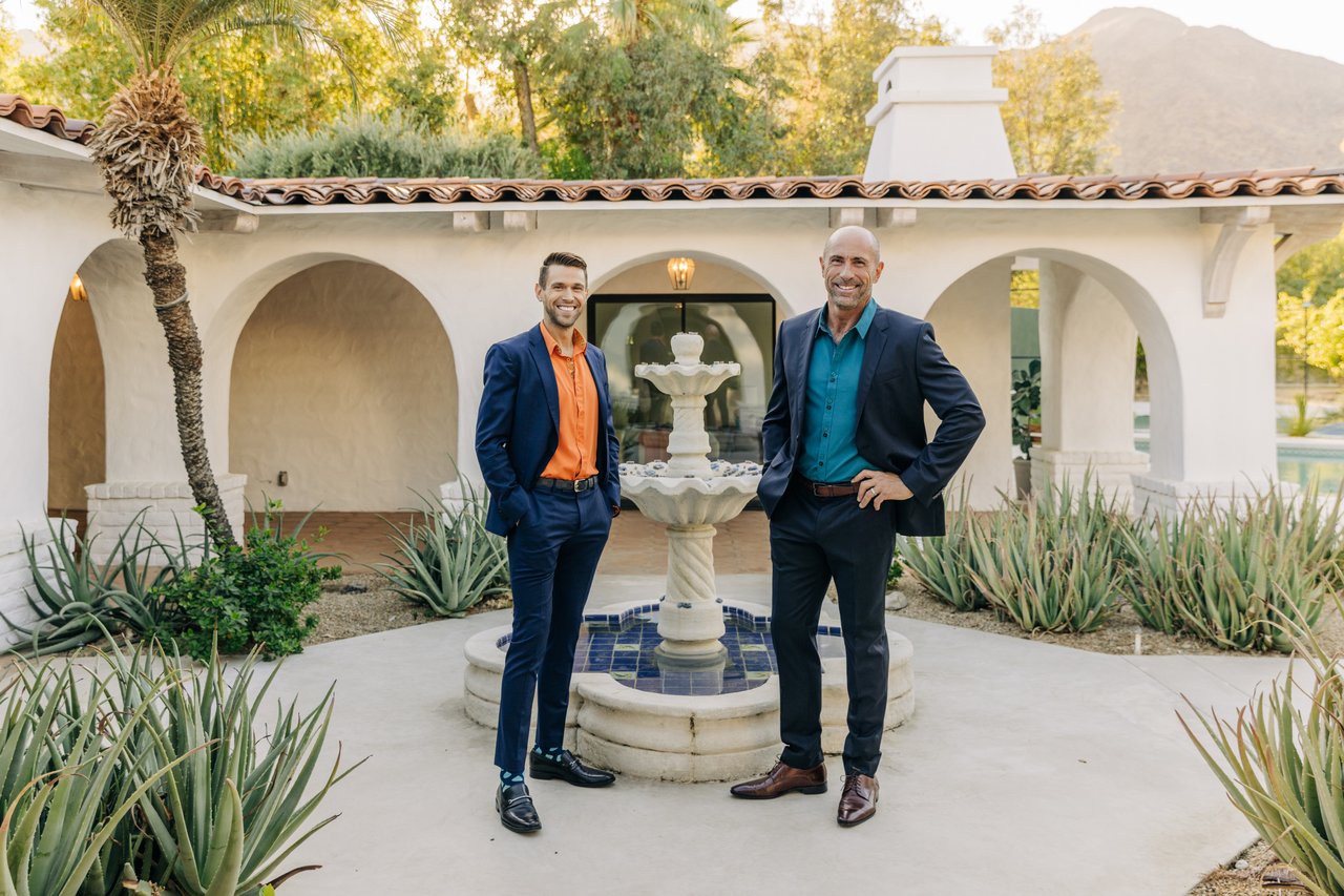 Two real estate agents pose beside a courtyard fountain in front of a white stucco home.