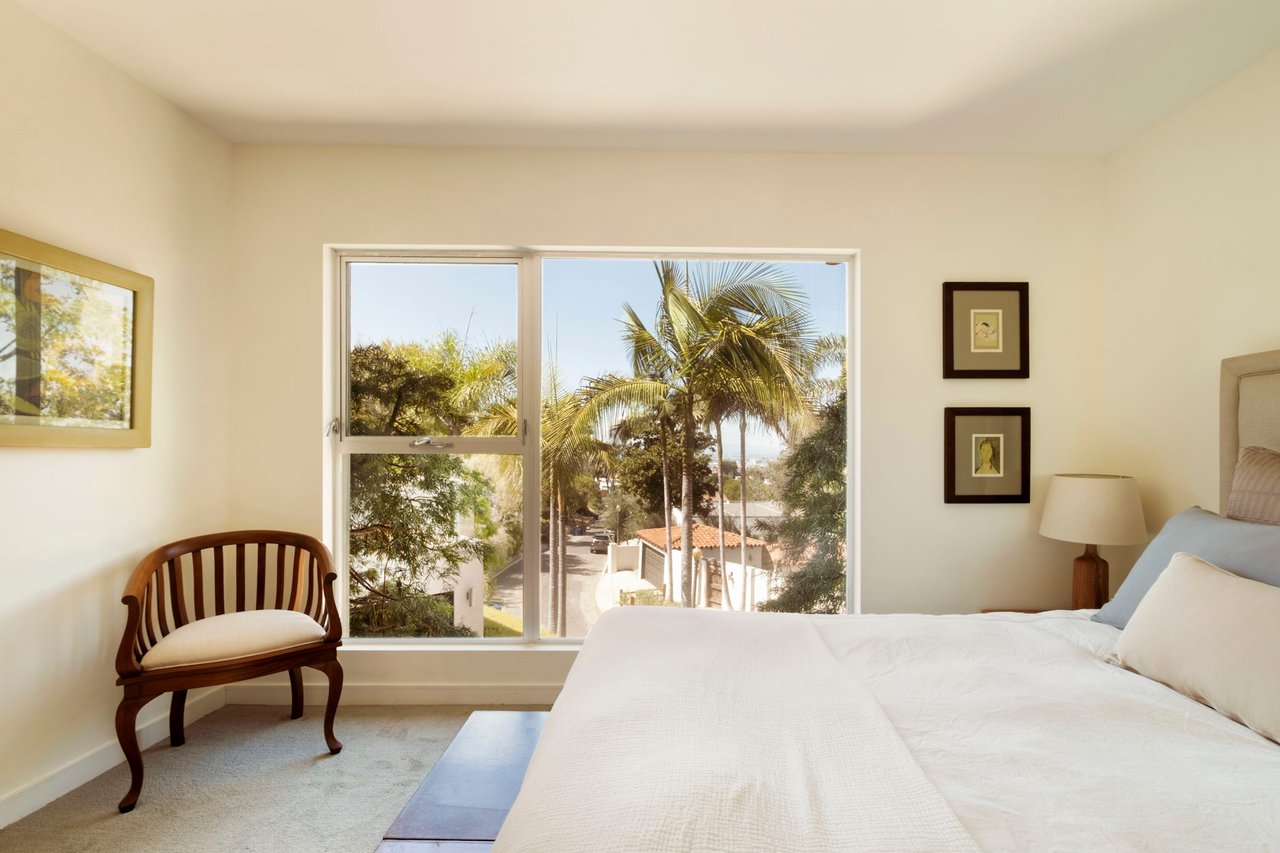 Bedroom with a white bed and wooden chair beside a large window overlooking palm trees.