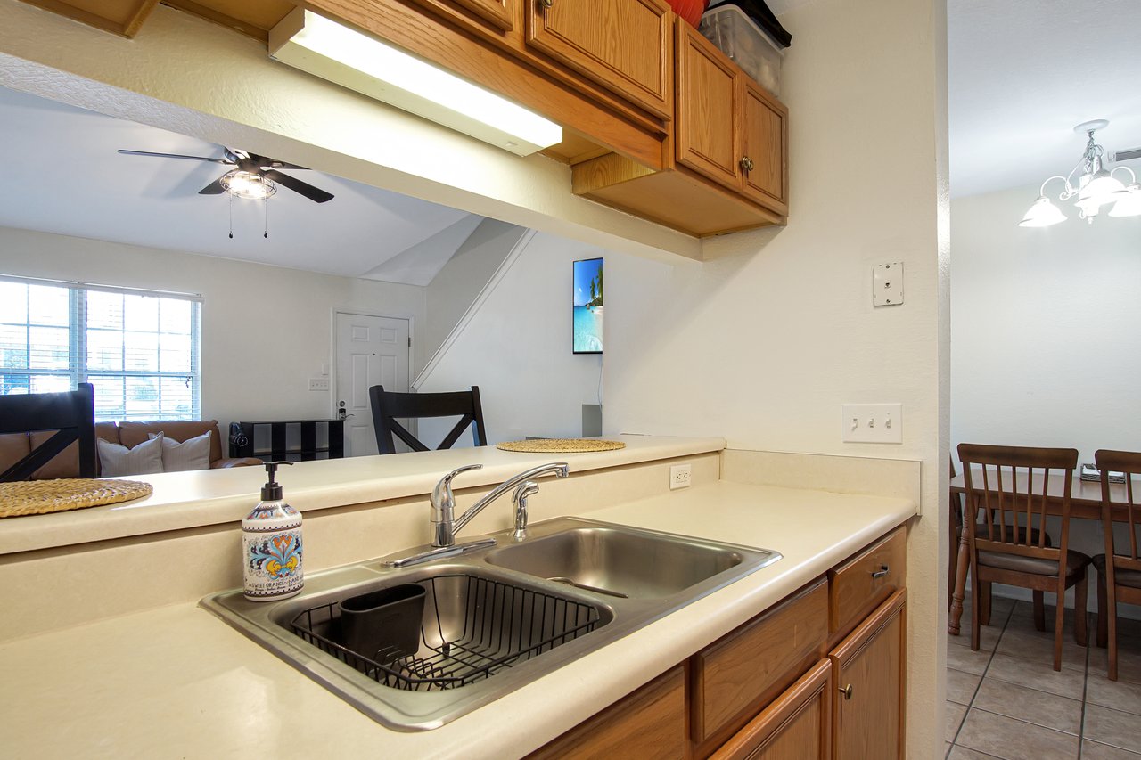 Bright kitchen with wooden cabinets and a beige countertop featuring a stainless steel sink. In the background, there's a cozy living area with a ceiling fan and a dining table with chairs, creating a welcoming atmosphere.
