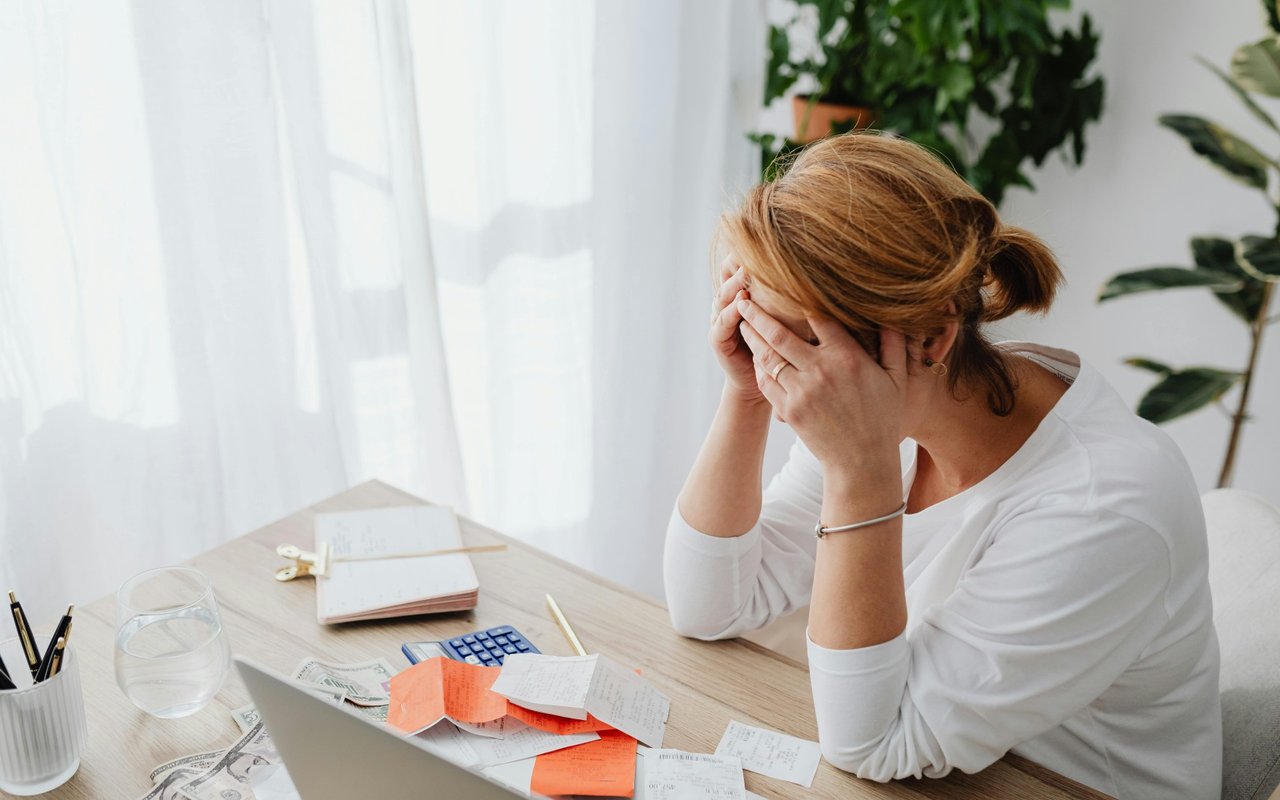 Woman covers her face at a desk scattered with bills, receipts, cash, and a calculator.