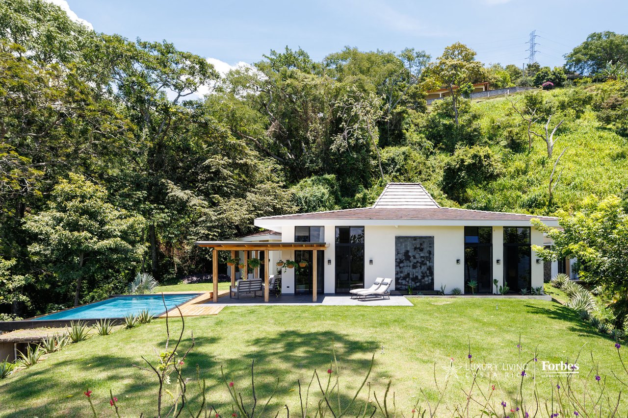 Modern white home with a covered patio and infinity pool amid lush greenery.
