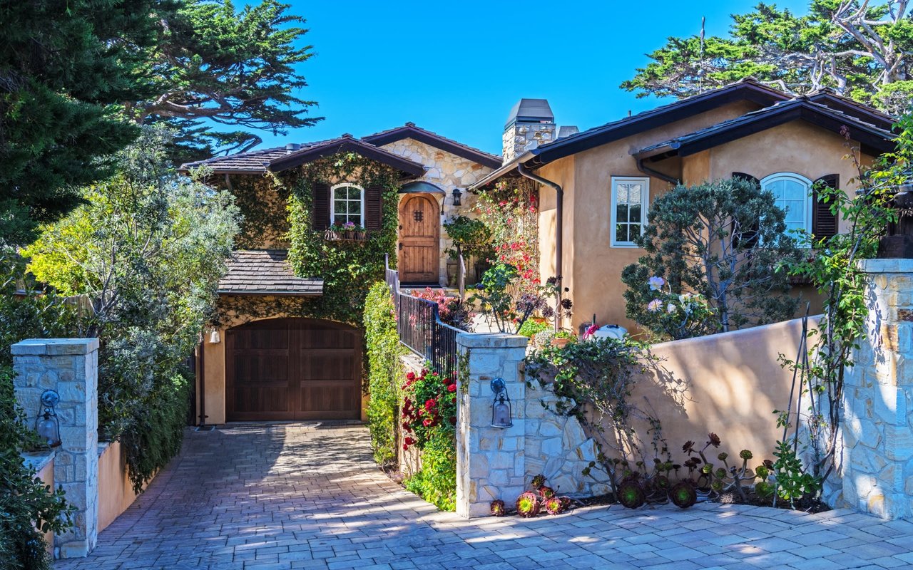 A charming, rustic-style house with a mix of light brown stucco and stone facades, and a dark shingle roof.