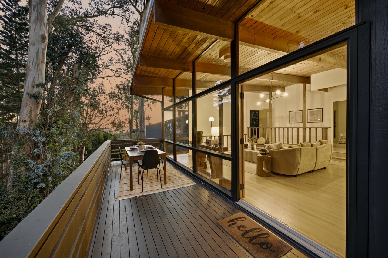 Wooden deck with outdoor dining set beside a glass-walled living room and wooded surroundings.