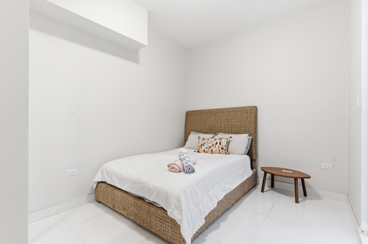 Bedroom with a woven tan bed frame, white bedding, and a small wooden side table.