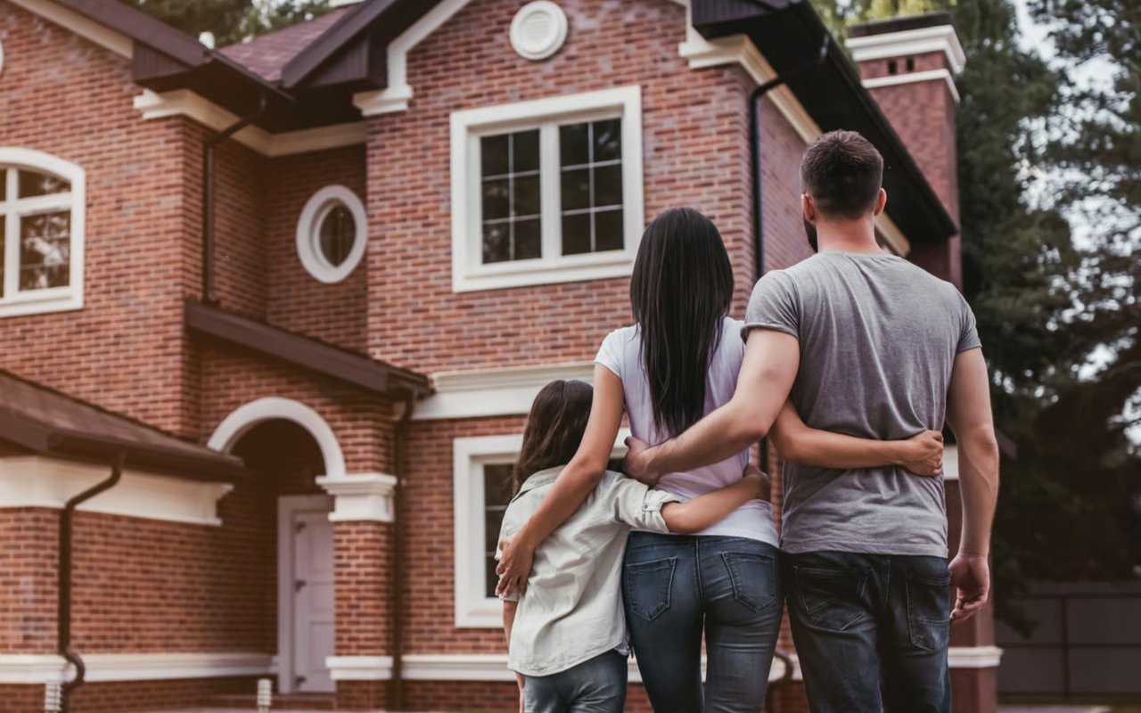 A family of three, with their backs to the camera, standing in front of a new brick house, with their arms around each other.