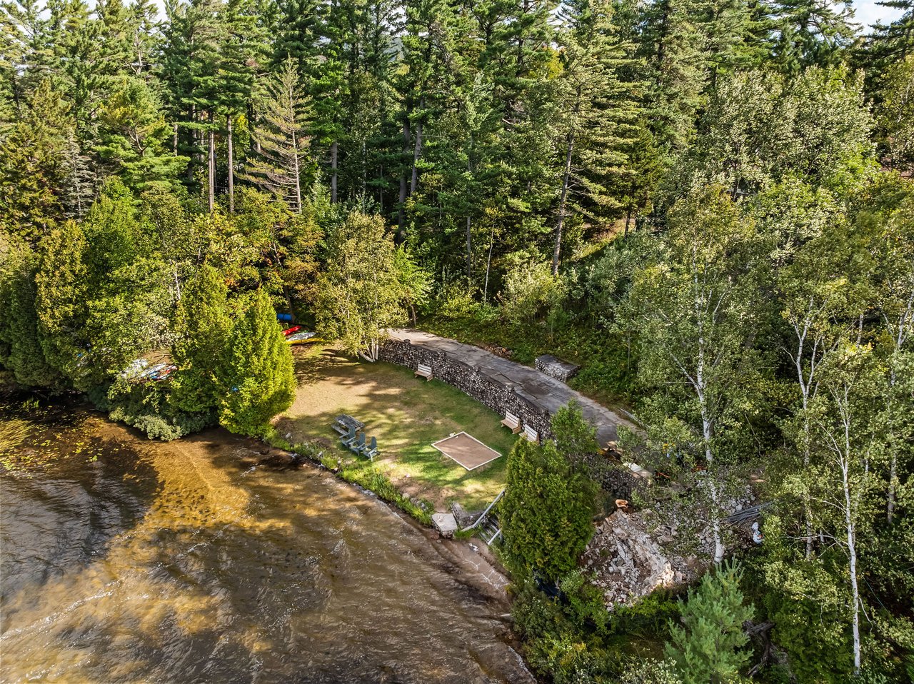 Wooded riverfront grounds with a grassy seating area, stone wall, and shallow water.