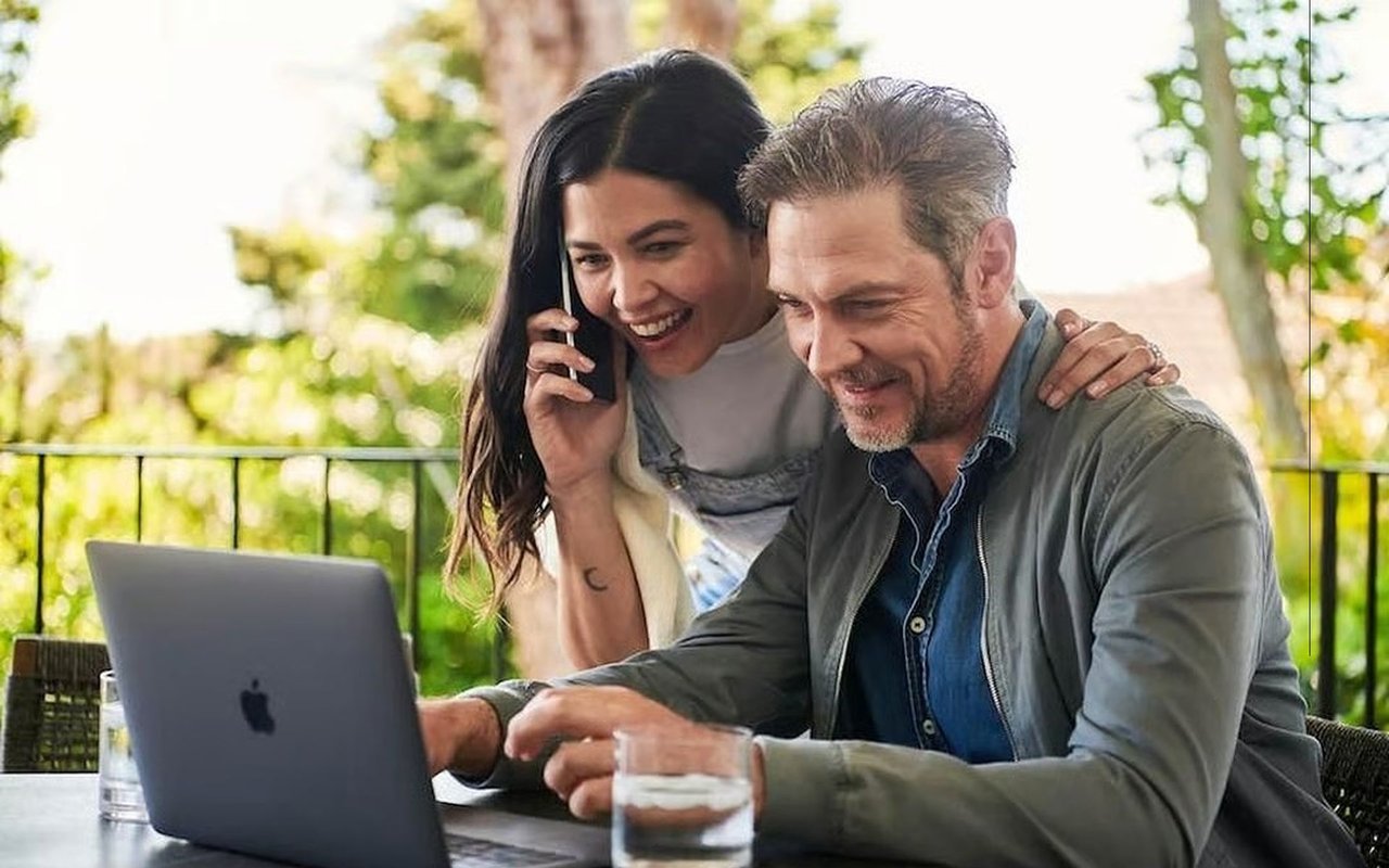 Couple reviews a laptop outdoors while the woman talks on her phone.