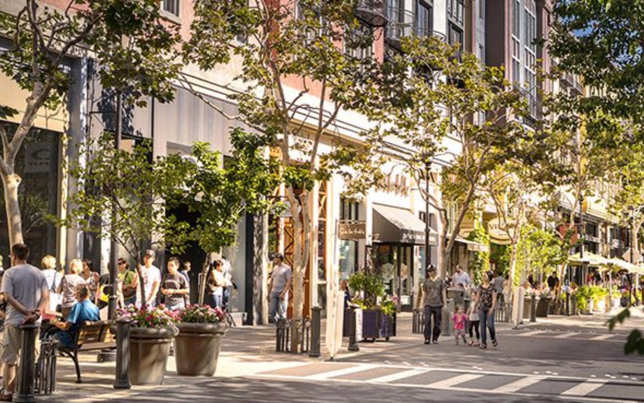 Pedestrians stroll past storefronts and leafy trees on a sunny shopping street.