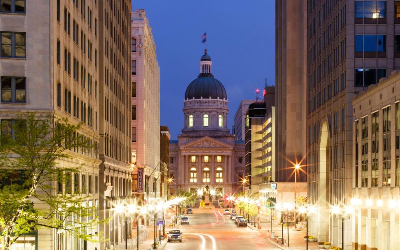 Downtown Indianapolis street at dusk, with the domed Indiana Statehouse between illuminated office buildings.