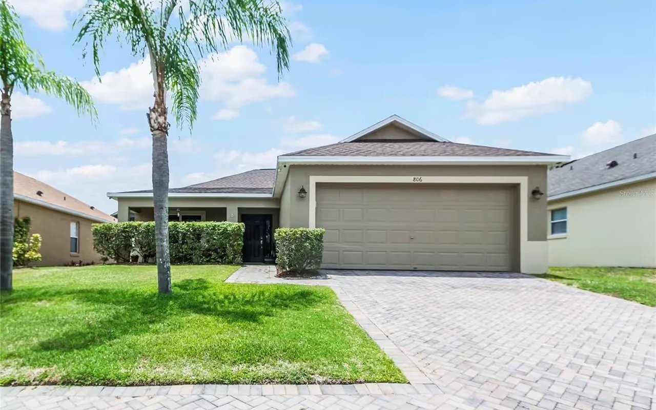 Single-story home with a two-car garage, paver driveway, trimmed hedges, and a palm tree.