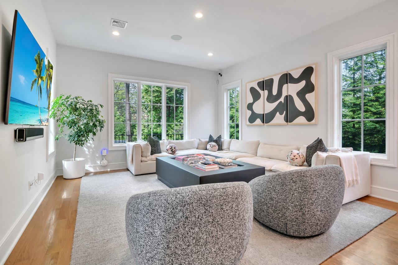 Bright living room with a white sectional, sculptural chairs, and large windows overlooking trees.