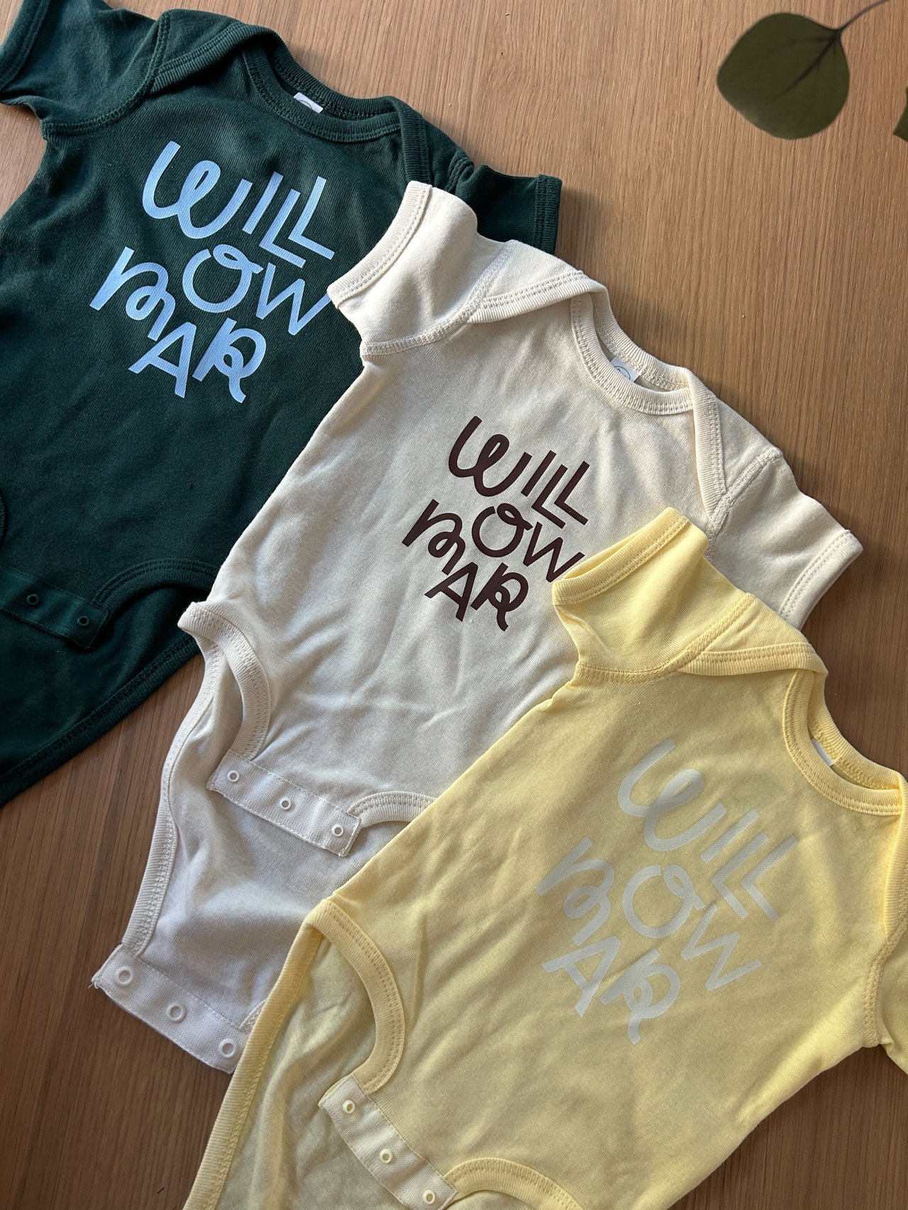 Three green, cream, and yellow baby bodysuits printed with Willowmar lettering lie on a wooden surface.