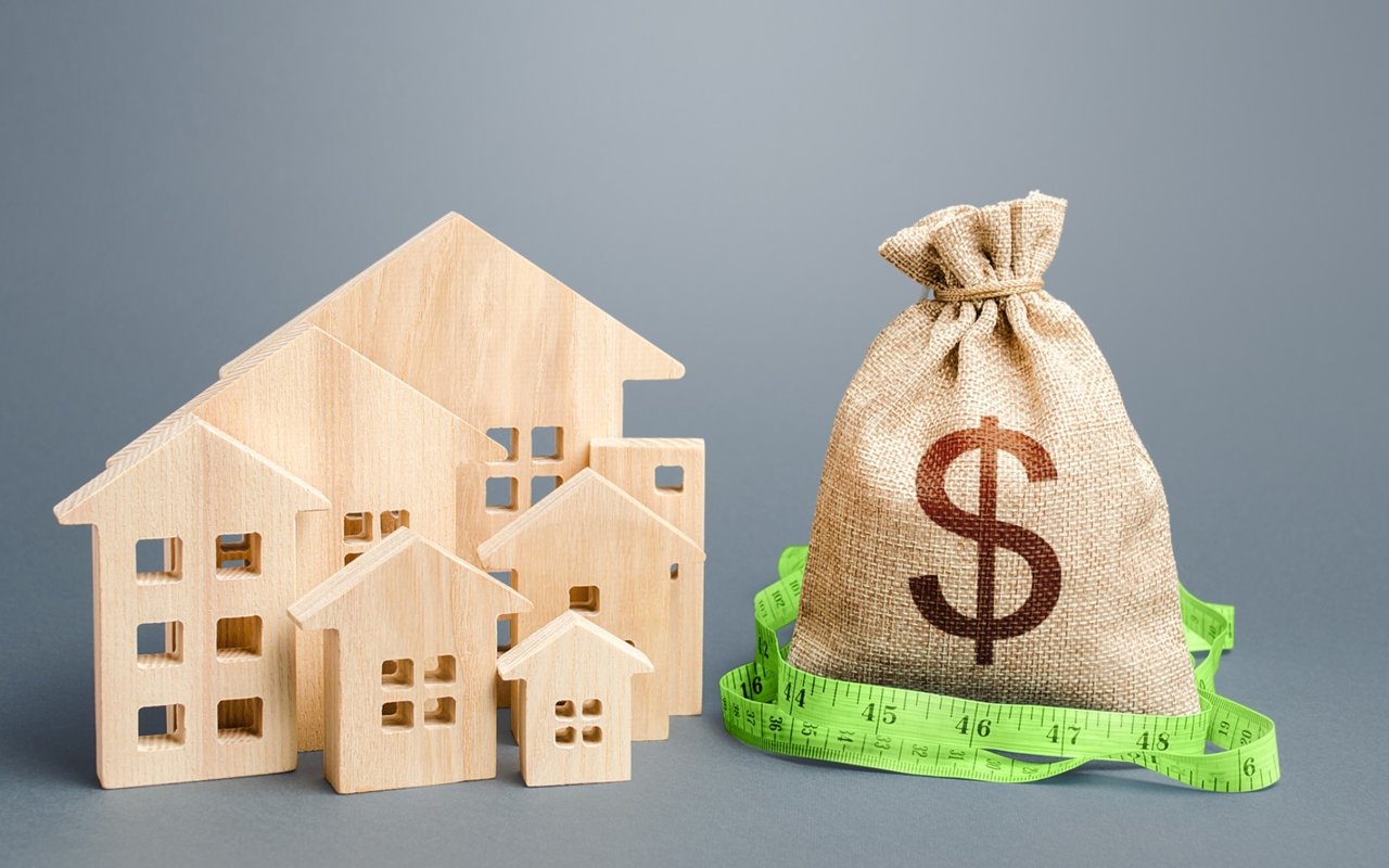 Wooden model houses beside a dollar-marked money bag wrapped in a green measuring tape.