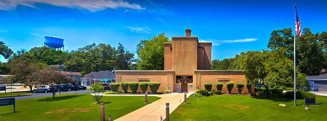 Brick schoolhouse with a tall central entrance tower, front lawn, and American flag.