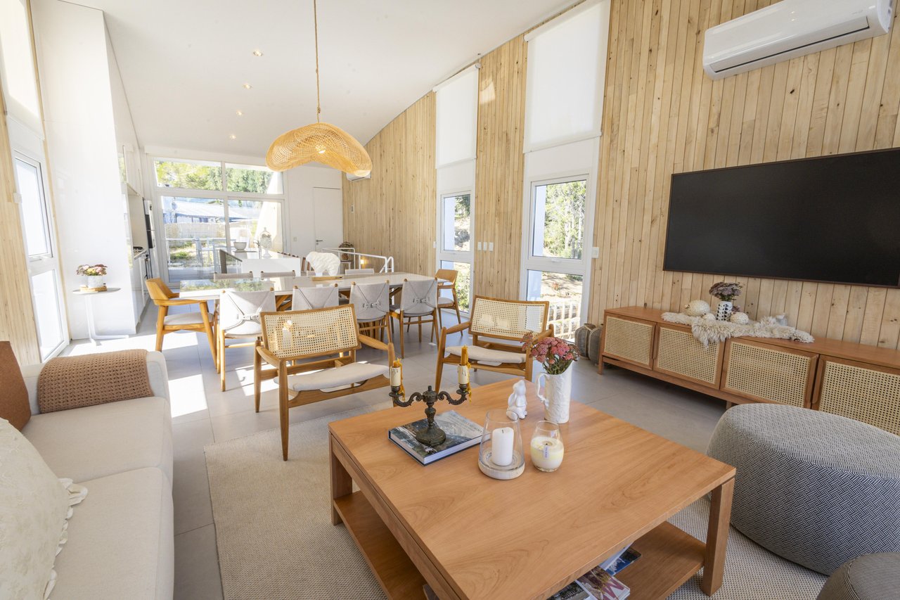 Living and dining area with light wood-paneled walls, large windows, and a wall-mounted television.