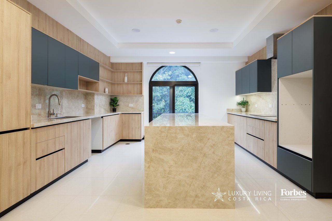 Modern kitchen with a large stone island, light wood cabinetry, and an arched window overlooking greenery.