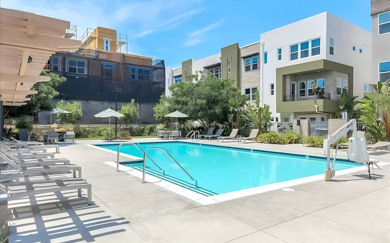  A community swimming pool with lounge chairs and umbrellas, surrounded by modern apartment buildings and lush landscaping.