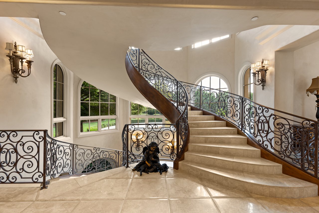 Grand foyer with a sweeping curved staircase, ornate iron railings, and tall arched windows.