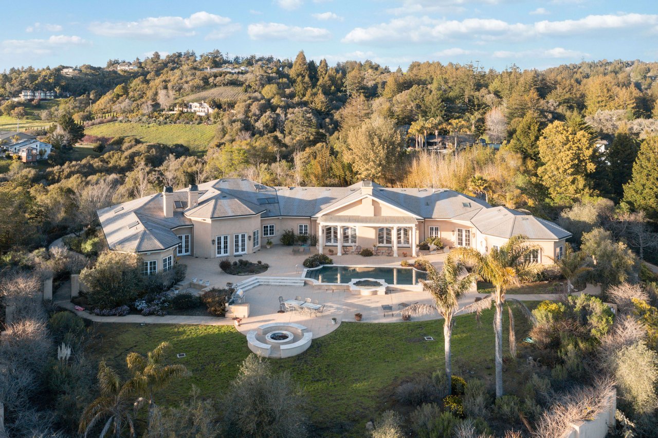 Aerial view of a sprawling home with a curved pool and circular fire feature, backed by wooded hills.