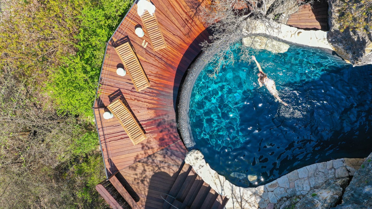 Overhead aerial of the grotto pool at Lick Creek Lodge, with its curved ipe sun deck and turquoise water set into the limestone, 4002 Tyx Trail, Spicewood, Texas.