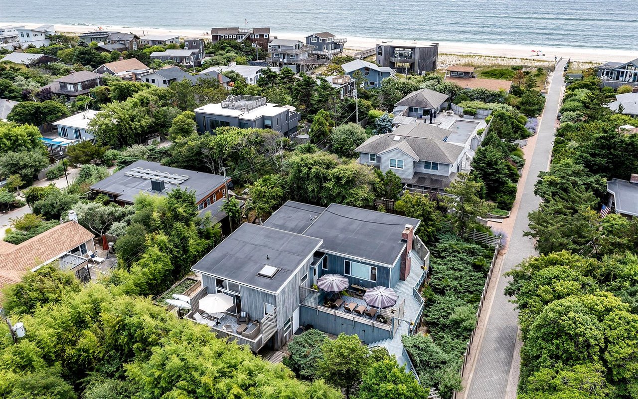 Aerial view of a gray beach house with broad decks, surrounded by coastal homes near the ocean.