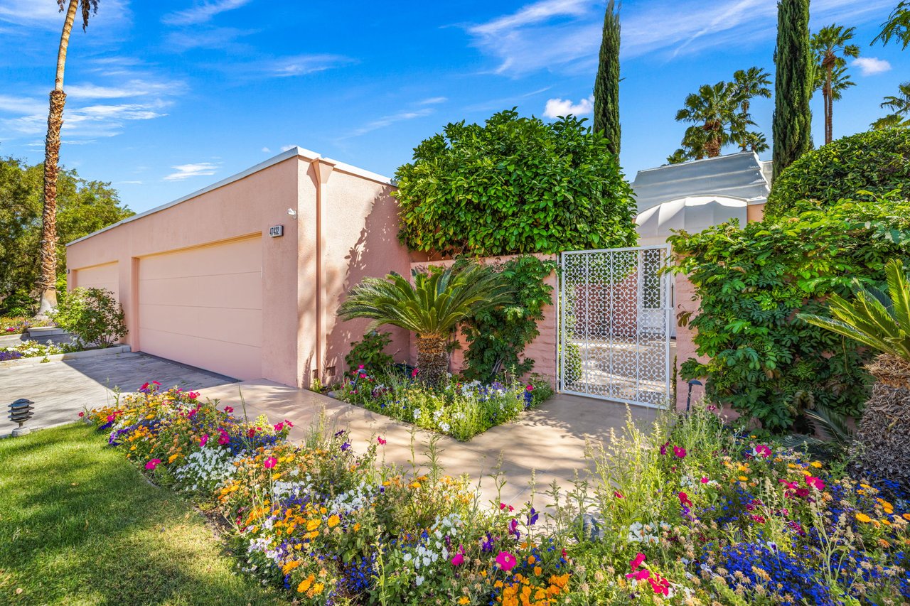 Pink stucco home with an ornate white gate, garage, and colorful flower beds.