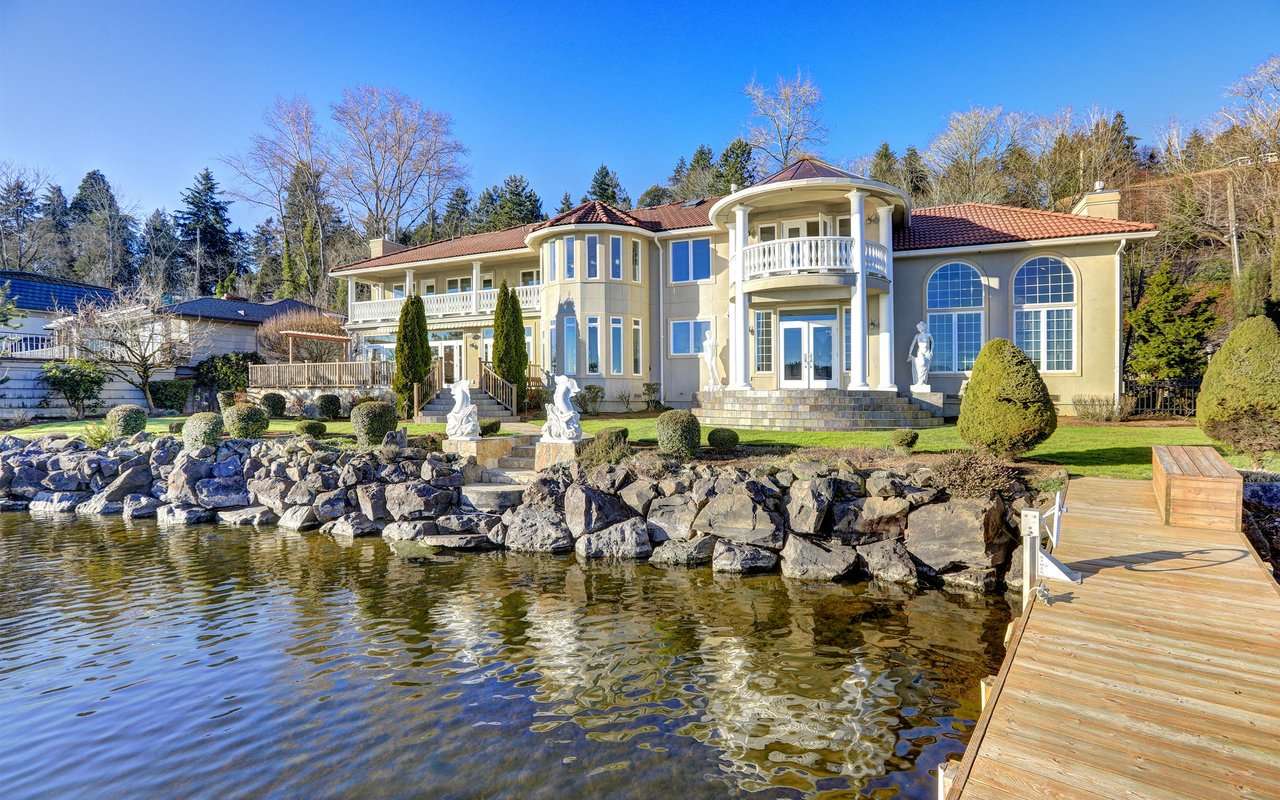 Large waterfront home with arched windows, balconies, rocky shoreline, and a wooden dock.