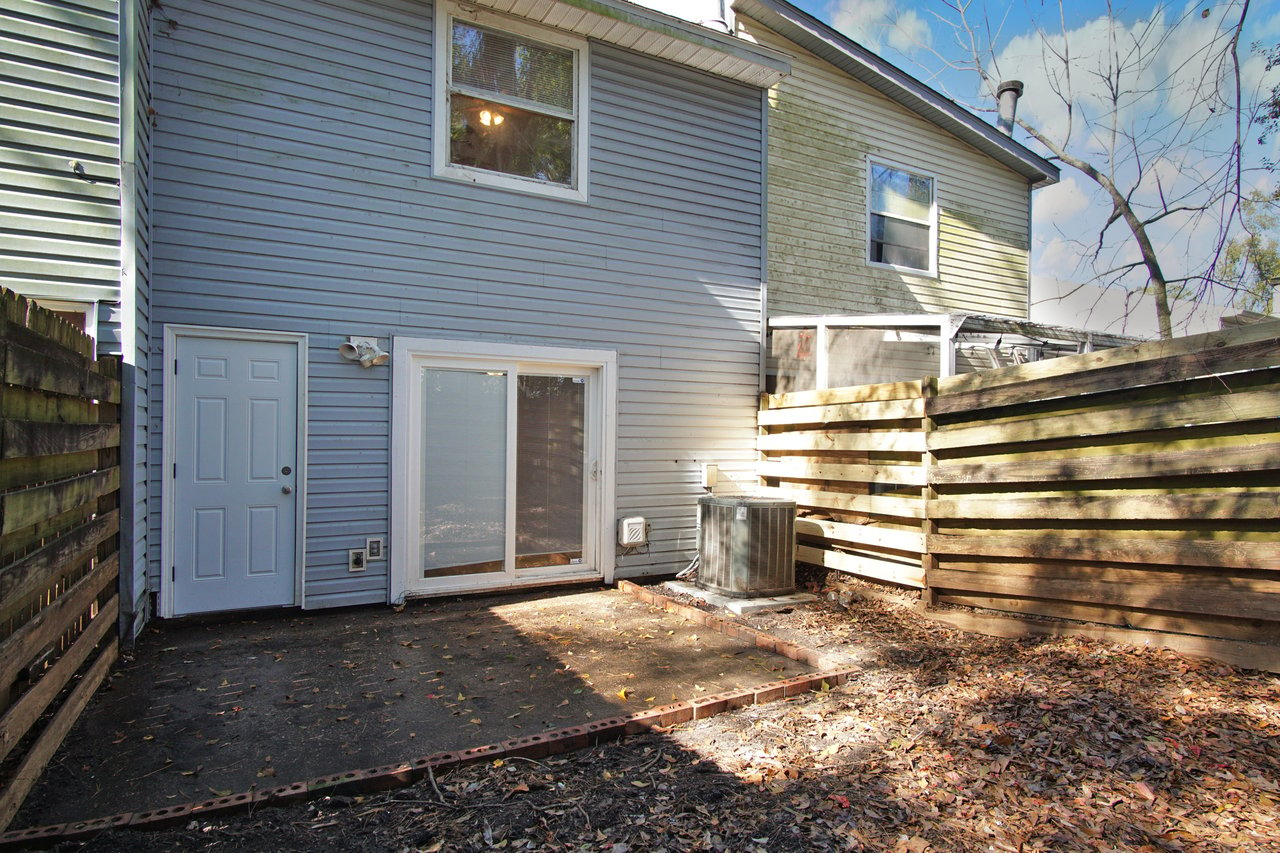 Small fenced backyard with a concrete patio area bordered by bricks. Blue siding house with a sliding glass door and utility unit. Sparse trees and fallen leaves.