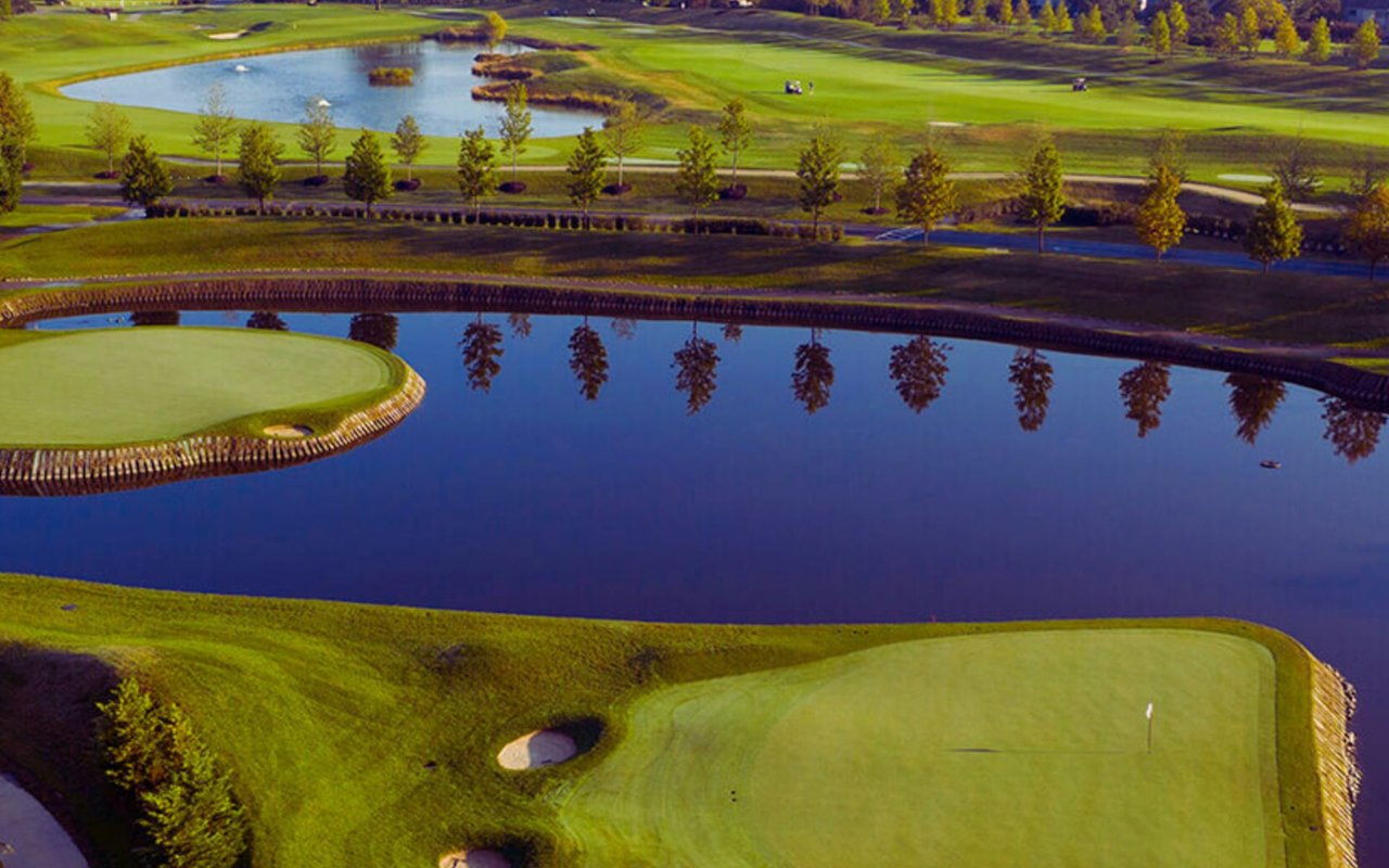 Golf course greens beside a large pond reflecting a row of trees.
