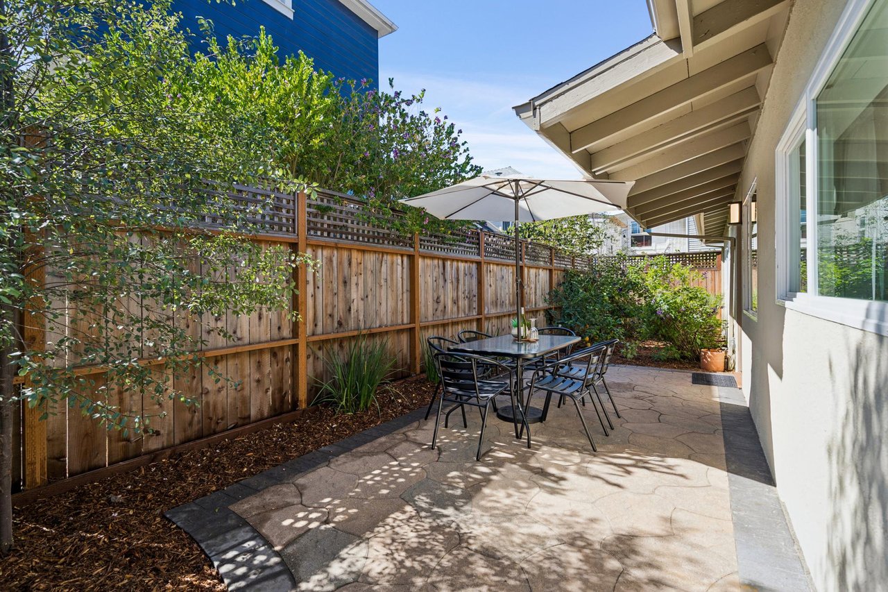 Stone-paved patio with umbrella-shaded dining set beside a landscaped garden and wooden fence.