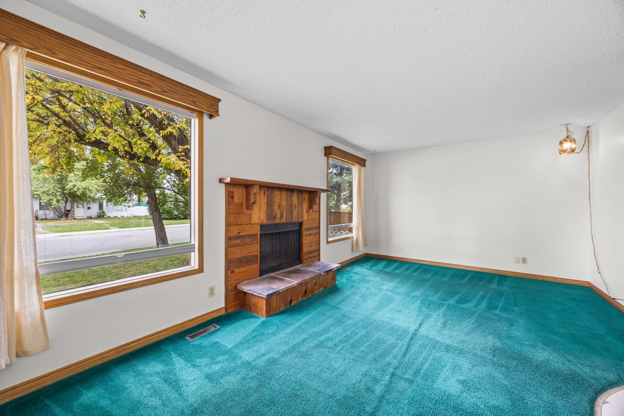Empty living room with turquoise carpet, a wood fireplace and large street-facing windows.