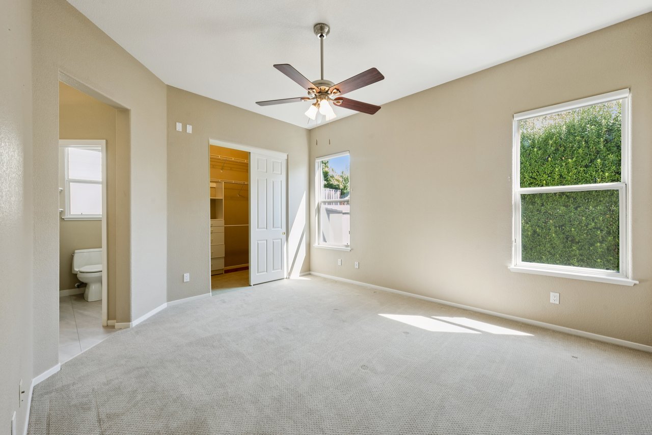 Empty bedroom with a ceiling fan, walk-in closet, adjoining bathroom, and windows overlooking a hedge.