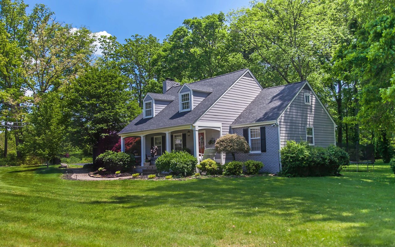 Gray-sided house with roof dormers and a covered porch, surrounded by mature trees and a broad lawn.