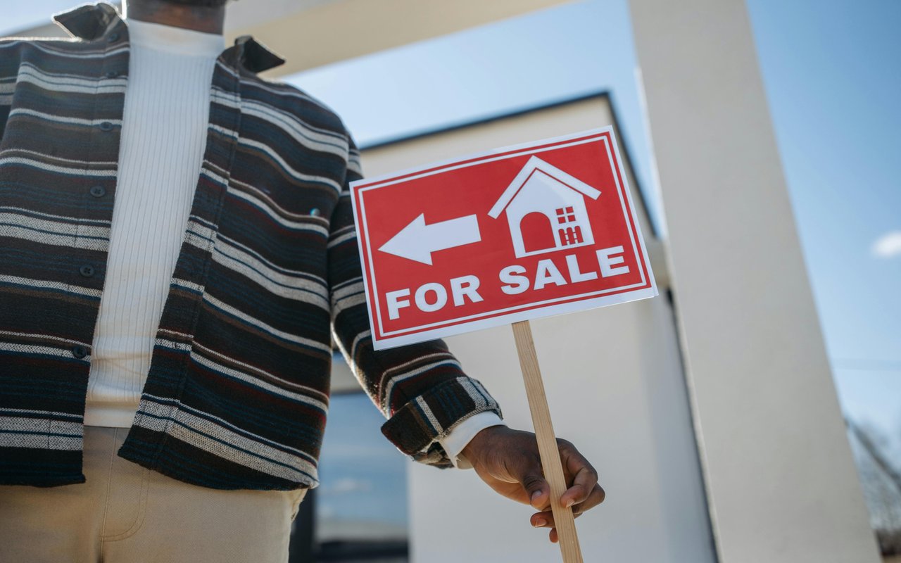 A person holds a red FOR SALE sign outside a modern building.