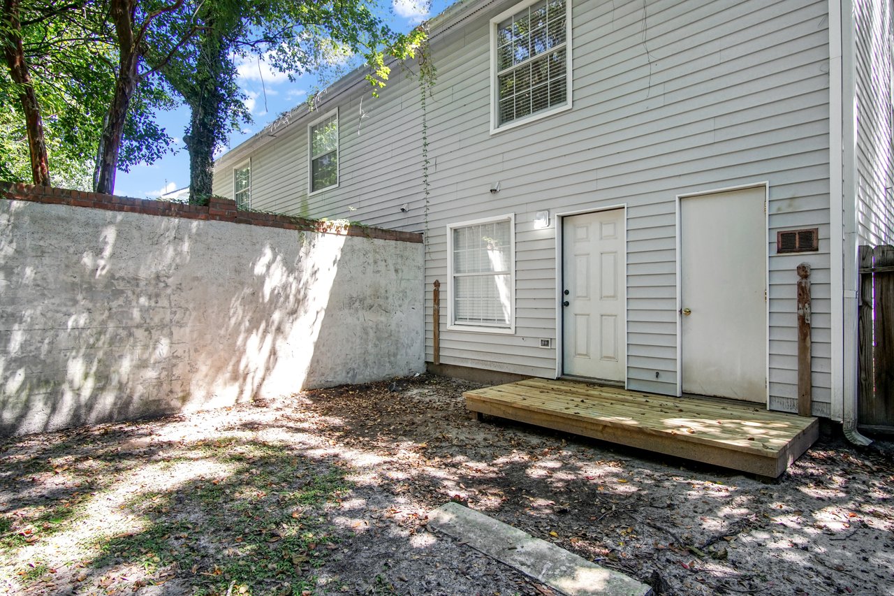 Backyard of 2506 Nugget Lane in Tallahassee, Florida, featuring a private fenced area with a tall stucco wall, mature trees providing shade, and a small wooden deck leading to the rear entry door of the home.