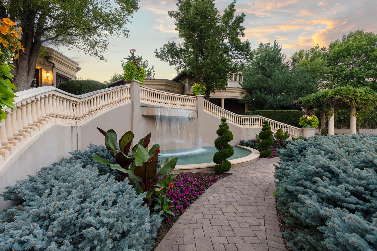 Estate garden with a tiered waterfall, curved walkway, flower beds, and sculpted shrubs at sunset.