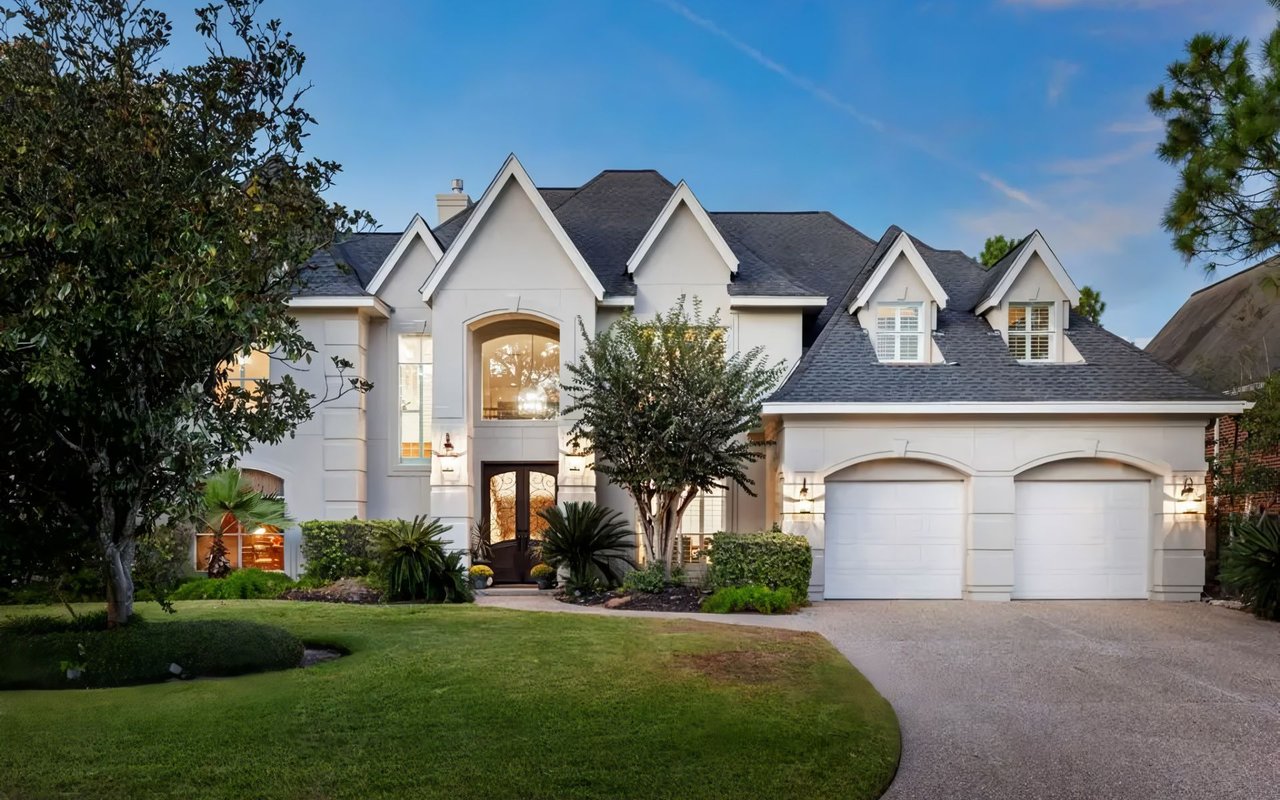  A large, stately two-story house with a light gray stucco exterior and a dark shingled roof stands prominently