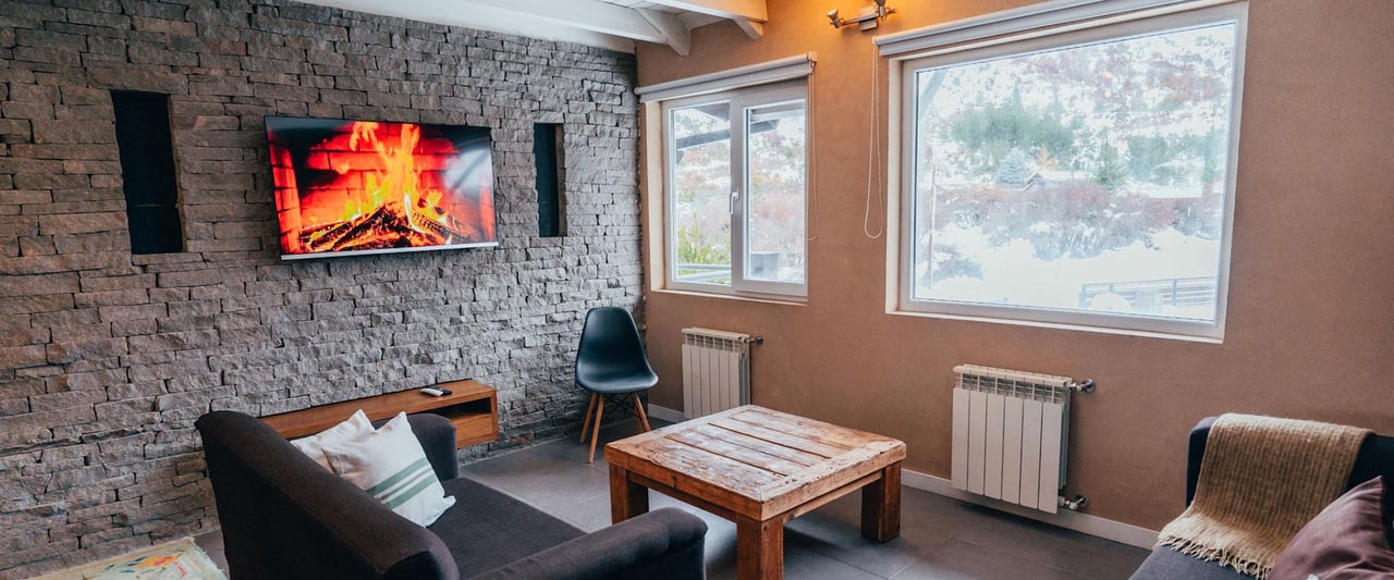 Living room with a stone accent wall, fire displayed on television, and windows overlooking snow.