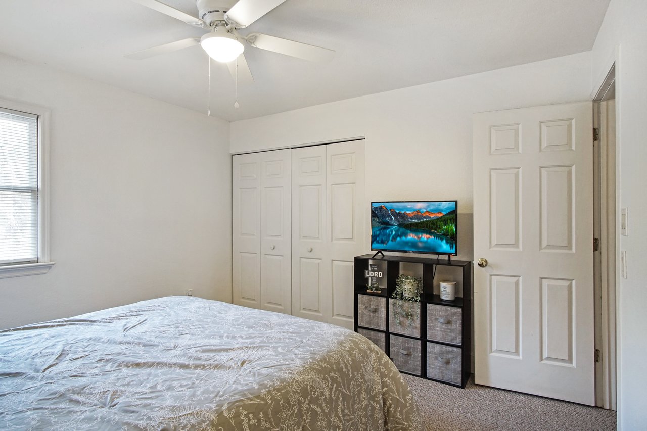 A cozy bedroom features a neatly made bed with a patterned duvet. A TV displays a scenic landscape on a cube shelf unit beside a white closet. Soft, warm lighting creates a calm atmosphere.