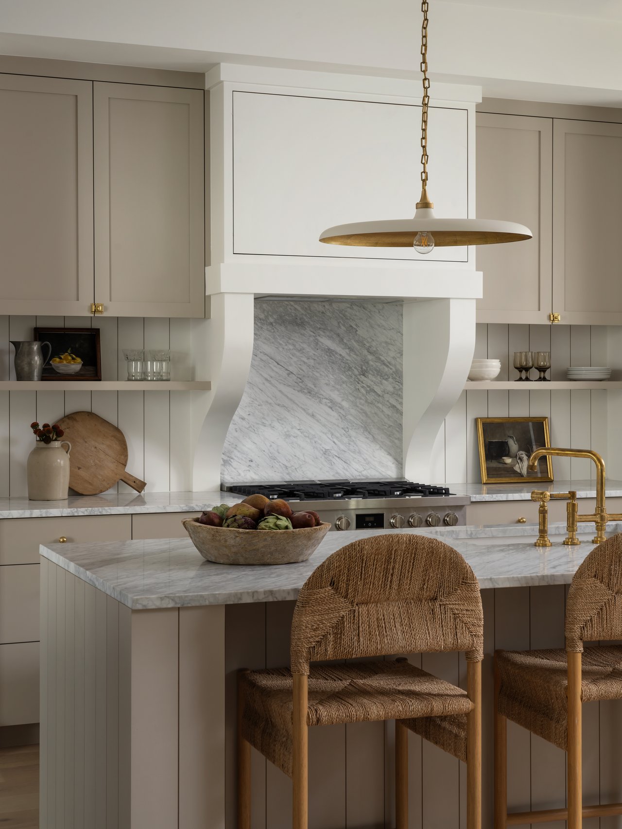 Kitchen with a marble range hood and island, brass fixtures, and woven bar stools.