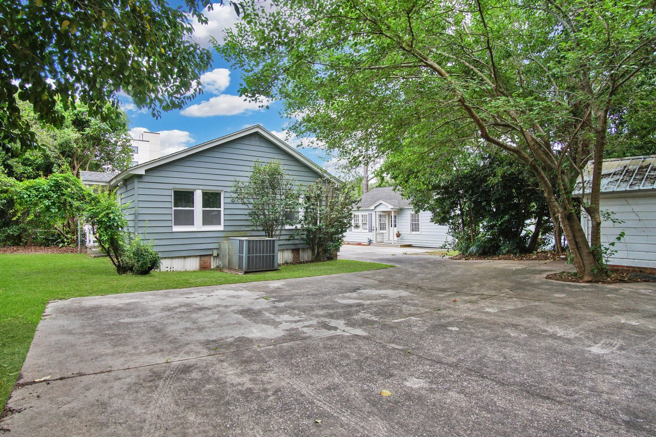 A small blue-gray house with white trim is surrounded by lush green trees and a neat lawn. A paved driveway leads to the entrance under a partly cloudy sky.