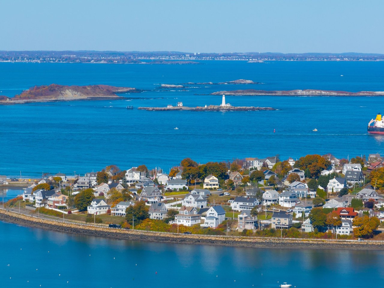 Aerial view of a coastal neighborhood beside blue water, with autumn foliage and a small offshore lighthouse.